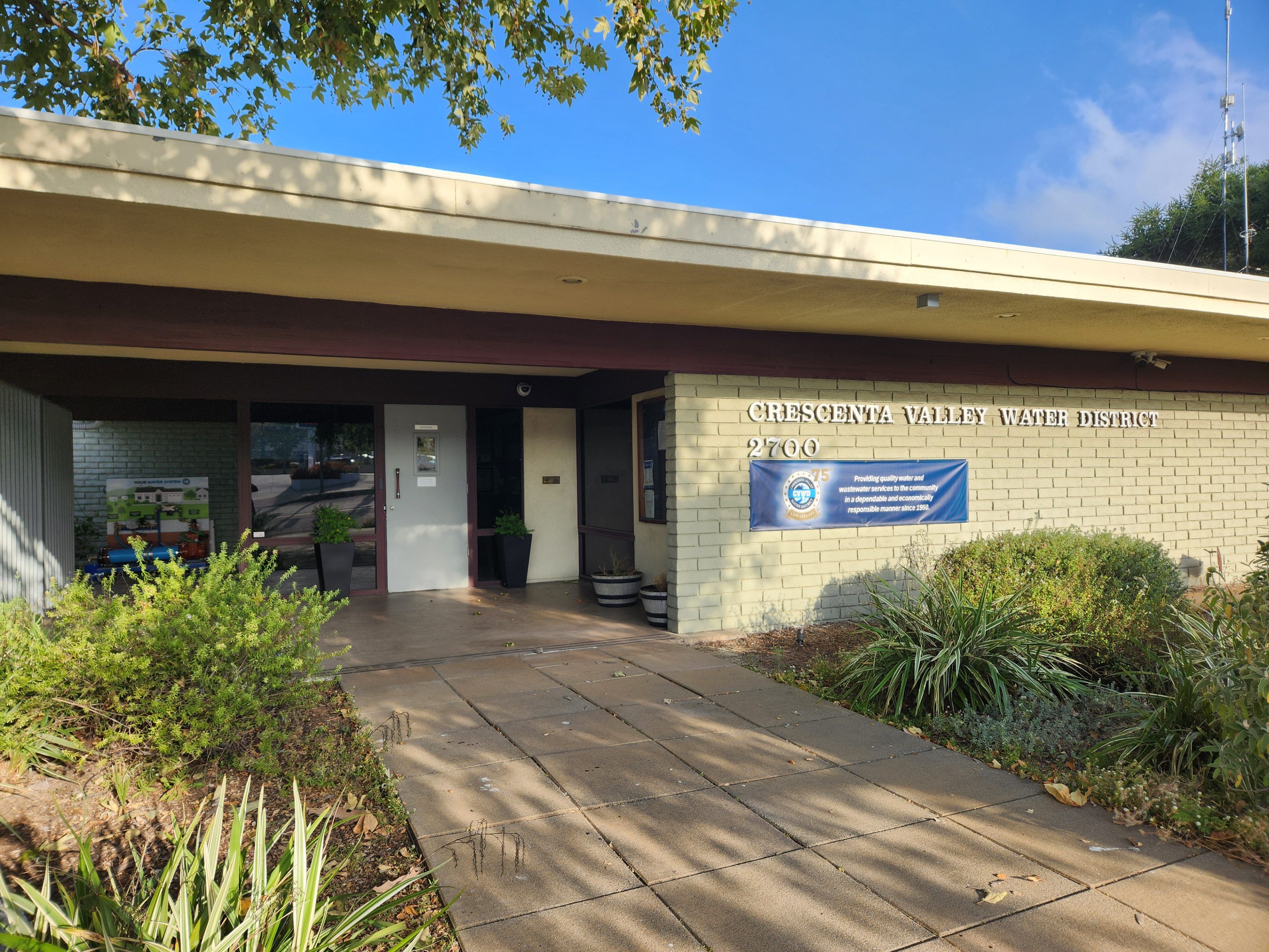 Crescenta Valley Water District building with a brick facade and blue banner. Plants and trees frame the entrance, creating a welcoming atmosphere.