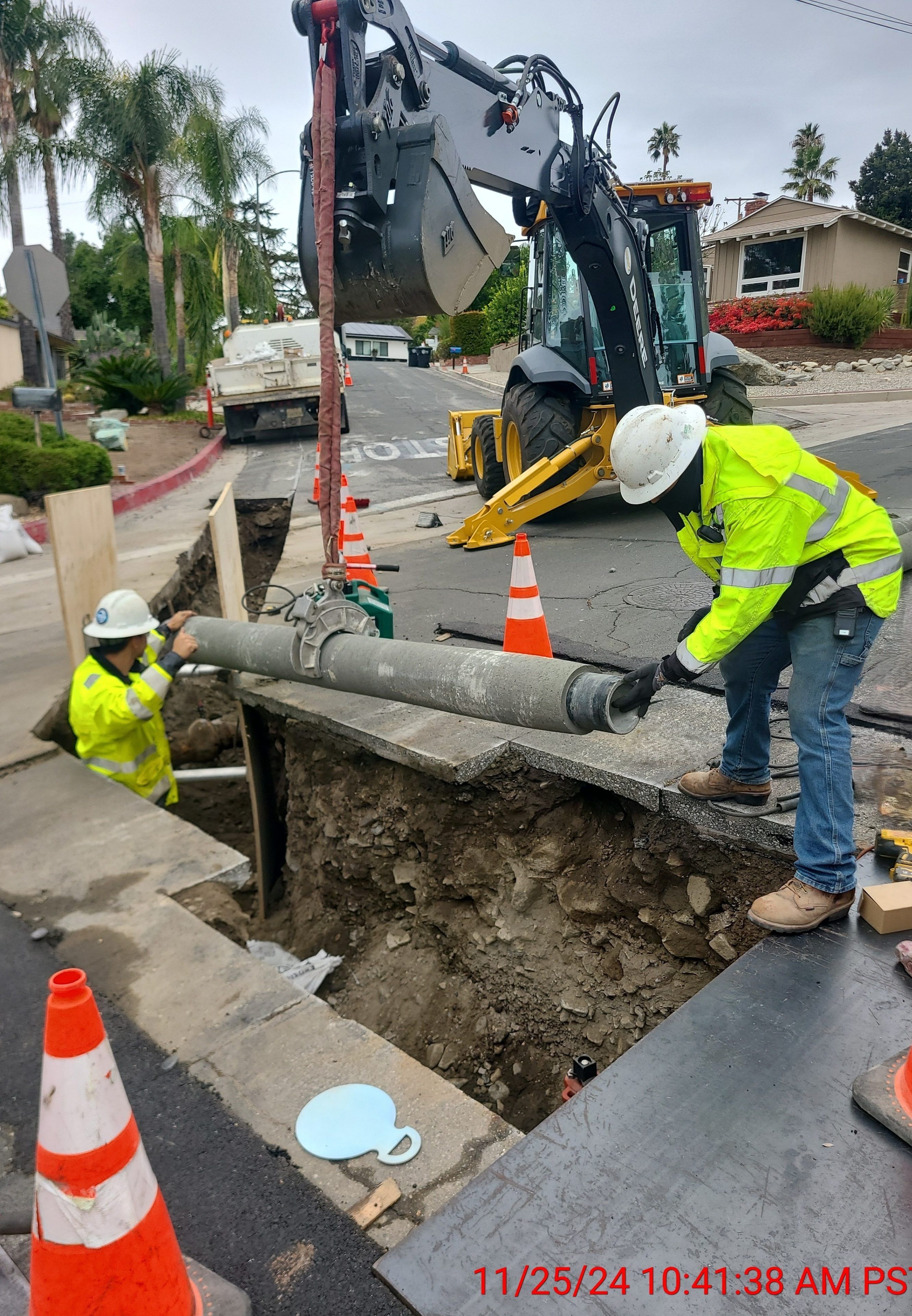 Construction workers in safety gear guide a metal pipe into a trench on a street. An excavator and traffic cones are visible. Cloudy day setting.