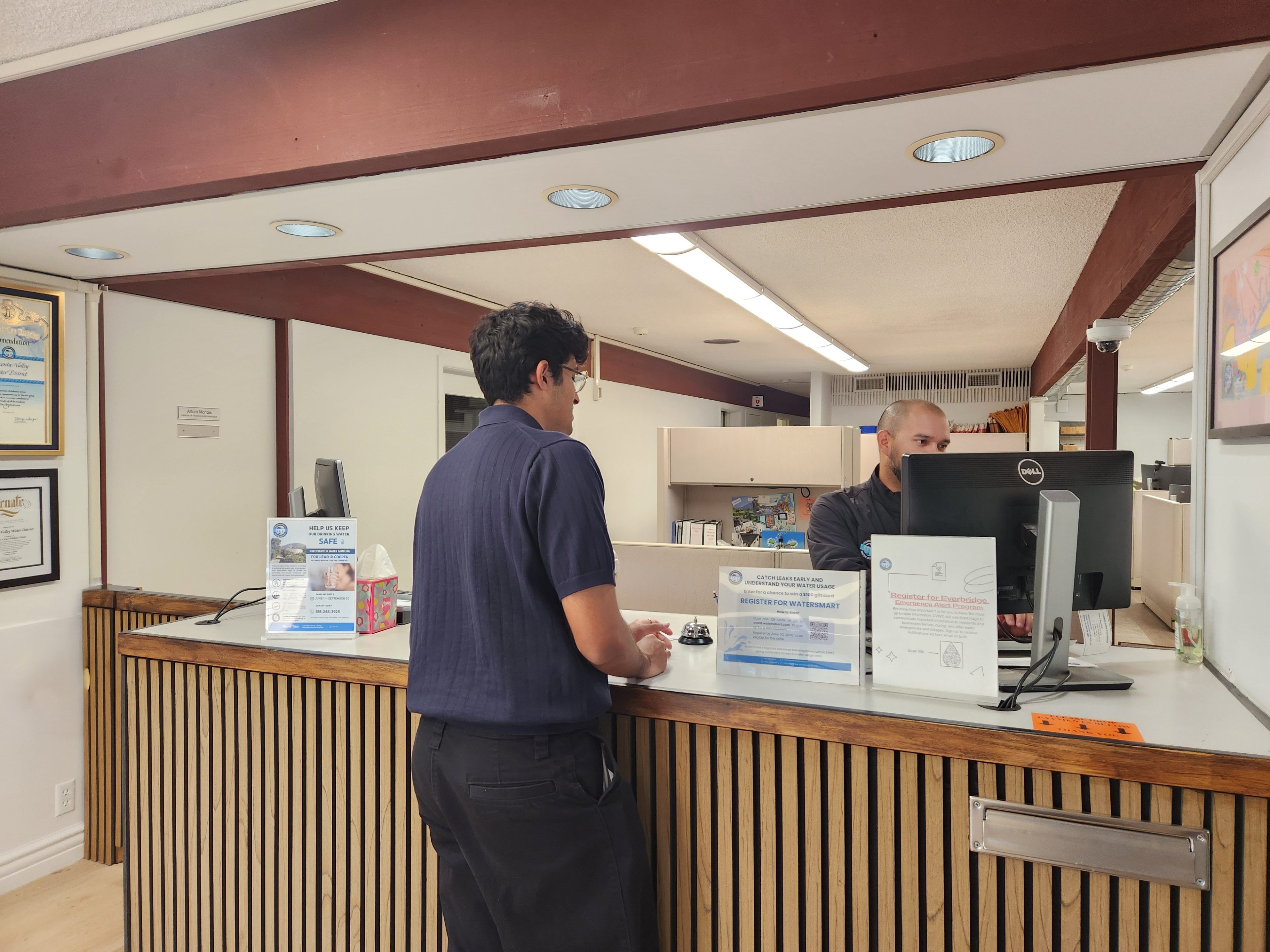 A man in a blue shirt speaks to a receptionist behind a wooden counter in an office. Informational signs and framed certificates are visible. The setting conveys a formal and professional tone.